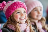 Young Caucasian girl wearing pink knit winter hat and scarf smiles joyfully with snow flakes on her clothing, capturing pure childhood happiness in cold season.
