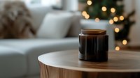 Cozy winter evening atmosphere with black scented candle in glass jar on wooden table, soft bokeh lights from Christmas tree in blurred background.