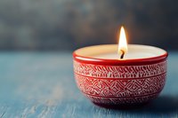Burning candle in red ceramic bowl with ethnic pattern on rustic blue wooden surface against dark background creates cozy atmospheric mood.
