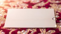 Blank white gift tag with eyelet on ornate red and gold patterned fabric background for holiday greeting cards and festive messaging.