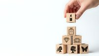 Hand placing wooden cube with crown symbol on pyramid of blocks featuring luxury icons including diamond, shell, and silhouettes on white background.