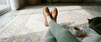 Cozy home relaxation scene with legs in green pants and brown loafers resting on a rug, a cup of tea nearby, and a tabby cat.