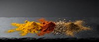 Colorful spice powders arranged in row on dark slate surface including turmeric, paprika, cumin and mixed seasonings for culinary cooking applications.