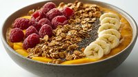 Healthy breakfast smoothie bowl with fresh raspberries, sliced banana, granola and chia seeds on orange base for nutritious morning meal preparation.