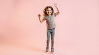 Happy young girl jumping with raised hands on pink studio background expressing joy and excitement.