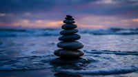 Balanced stone stack on ocean shore during sunset with purple and pink sky colors creating peaceful meditation scene for wellness and mindfulness concepts.