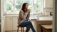 Thoughtful Caucasian middle-aged woman sitting in bright kitchen with coffee cup and laptop, working from home in cozy casual style.
