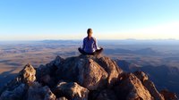 Young woman in athletic wear meditating in lotus position on mountain peak at sunrise, overlooking vast desert landscape and distant mountain ranges in peaceful solitude.