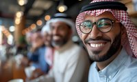 Cheerful Middle Eastern businessman wearing traditional keffiyeh and glasses smiling during corporate meeting, with colleagues in background. Positive workplace atmosphere.