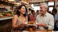 Mature couple enjoying meal together at busy restaurant, sharing food and laughing. Happy dining experience with warm atmosphere and social gathering.