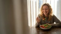 Happy blonde woman enjoying fresh healthy salad at wooden table by window in bright modern kitchen for wellness lifestyle and nutrition concepts.