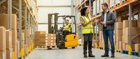 Warehouse workers discussing logistics while forklift operator manages inventory in modern distribution center with organized shelving systems.
