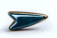 Stylized metallic arrow pin with teal blue enamel finish and gold trim captured in macro photography against white background, showing fine detail and reflection.