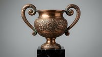 Ornate bronze trophy cup with decorative handles and floral engravings on marble base for awards ceremonies and competitions.