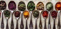 Colorful array of dried herbs and spices in vintage silver spoons arranged in row on rustic background, including turmeric, paprika, and mixed dried flowers for cooking.