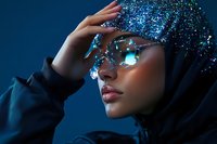 Young woman in hijab wearing futuristic crystal glasses and glitter head covering against deep blue background, creating modern fashion portrait.