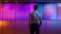 Professional in business attire observing financial data on large digital display with vibrant purple and orange gradient lighting, showing market analysis and trading charts.