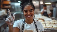 Young African American female baker showing peace sign while smiling in commercial kitchen, wearing apron with flour-covered hands and fresh bread dough in background.