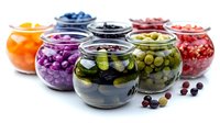 Colorful assortment of pickled vegetables and fruits in glass jars, including purple cabbage, cucumbers, olives, berries and citrus on white background.