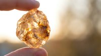Hand holding translucent amber gemstone with natural inclusions against warm sunlight creating golden glow and bokeh background for jewelry design.
