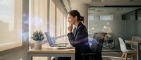 Young professional businesswoman working on laptop in modern office space with natural sunlight streaming through windows during productive workday.