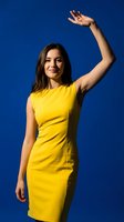 Young Hispanic woman in bright yellow dress waving hand against vibrant blue background. Cheerful greeting gesture for marketing and communication concepts.