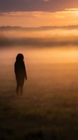 Silhouette of woman standing in misty field during golden sunrise with warm orange sky and atmospheric fog creating peaceful morning landscape scene.