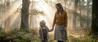 Young Caucasian mother and daughter holding hands in misty autumn forest with sunbeams, warm family bond in nature, peaceful outdoor moment.