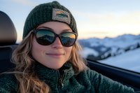 Young woman in winter attire with sunglasses and knit beanie smiling during sunset in snowy mountain landscape, capturing carefree outdoor adventure lifestyle.