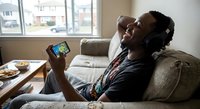 Young African American man relaxing on couch with headphones and smartphone, enjoying music and mobile entertainment at home during leisure time.