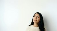 Young woman with long dark hair and South Asian ethnicity wearing a cream knit sweater, looking upward thoughtfully against a white wall background for portrait and lifestyle use.