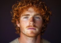 Close up portrait of young man with bright ginger curly hair, blue eyes and freckles against dark background, showing natural masculine features and confidence.