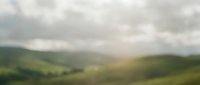 Soft-focused rolling green hills under cloudy sky with golden sunlight breaking through clouds, creating serene countryside landscape for nature and travel projects.