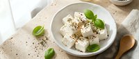 Fresh white cheese cubes with herbs and basil leaves in ceramic bowl on rustic wooden table, healthy Mediterranean appetizer ready for cooking.