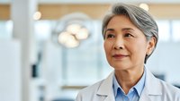 Confident mature Asian female doctor in white coat smiling warmly in modern medical facility, representing healthcare professionalism and patient care.