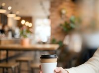Closeup of hand holding white takeaway coffee cup with black lid in modern cafe interior with blurred background and warm lighting for beverage and lifestyle use.