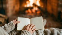 Unrecognizable person in cozy sweater holding open book near fireplace, enjoying reading by warm fire during winter relaxation and leisure time.