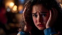 Young woman with curly hair expressing worry and anxiety in dark atmospheric lighting with bokeh background, hands on face showing distress and concern.