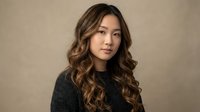 Professional Asian woman with wavy brown hair wearing dark sweater against neutral background for business and lifestyle content.