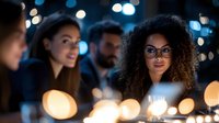 Young African American woman with curly hair and glasses illuminated by warm bokeh lights at evening gathering, surrounded by diverse friends sharing conversation.