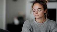 Young Caucasian woman with closed eyes and slight smile wearing gray knit sweater, expressing serenity and mindfulness in soft natural lighting against blurred background.