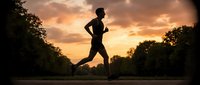 Male athlete jogging on rural road at golden sunset with trees, silhouetted runner training outdoors for fitness, marathon preparation, and healthy lifestyle.