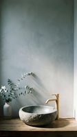 Minimalist bathroom vanity with round stone vessel sink, brass faucet, and eucalyptus stems in ceramic vase against textured gray wall for spa and interior design projects.