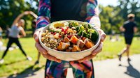 Person holding nutritious bowl with quinoa, roasted vegetables and greens outdoors during community fitness event in park setting.
