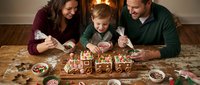 Young family decorating gingerbread house together during holiday season. Parents and child enjoying festive baking activity at home with candies and icing.