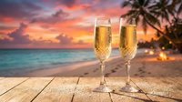 Two champagne flutes filled with golden bubbly on wooden deck overlooking tropical beach at sunset. Palm trees frame romantic ocean view for celebration.