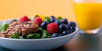 Grilled chicken breast with fresh berries and spinach on white plate, orange juice glass in background. Healthy protein-rich meal for clean eating lifestyle.