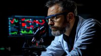 Bearded scientist in lab coat and glasses conducts research using microscope against background of digital data screens showing market analysis graphs.