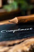 Handwritten word compliment in white chalk on dark surface with wooden pencil in soft focus background, creating inspirational atmosphere for greeting cards.