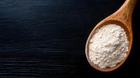 White flour in wooden spoon on dark black wooden background, top view. Minimalist food photography composition with copy space for baking and cooking.